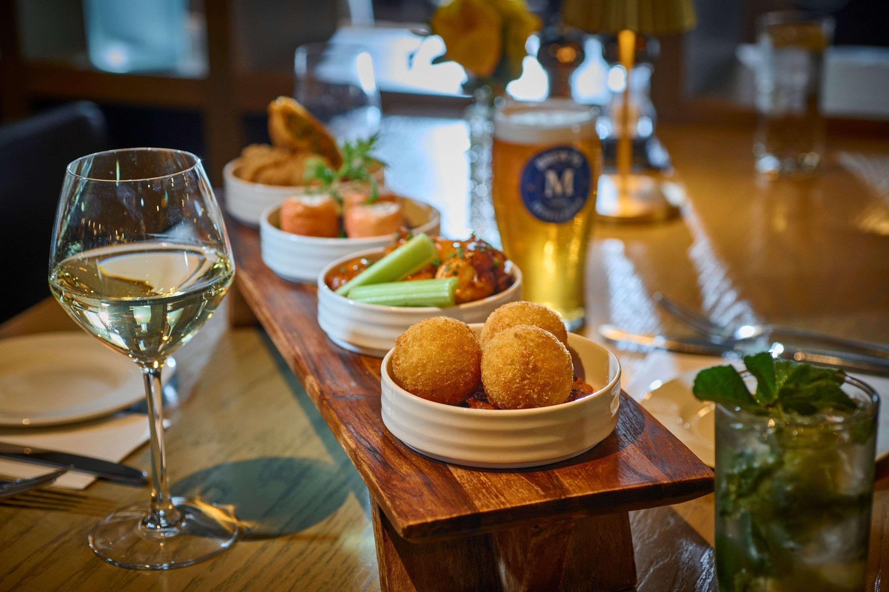 Una mesa de comedor con una copa de vino blanco, un bol de croquetas, una jarra de cerveza y un vaso de bebida helada.