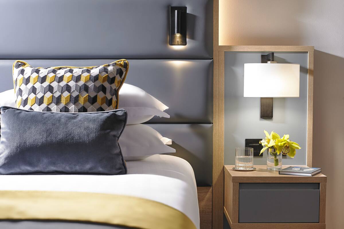 A neatly arranged hotel room with a bed, pillows, a bedside table with a lamp, a vase with flowers, and a framed picture on the wall.