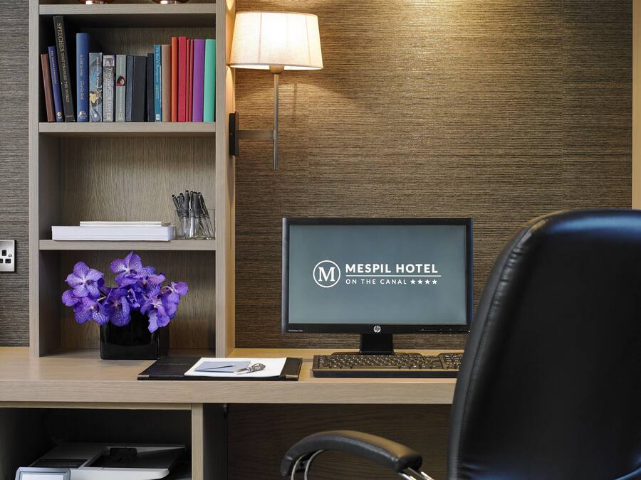 Una recepción de hotel con un monitor de computadora mostrando el nombre del hotel, una lámpara de escritorio, un jarrón con orquídeas moradas y una estantería con varios libros.
