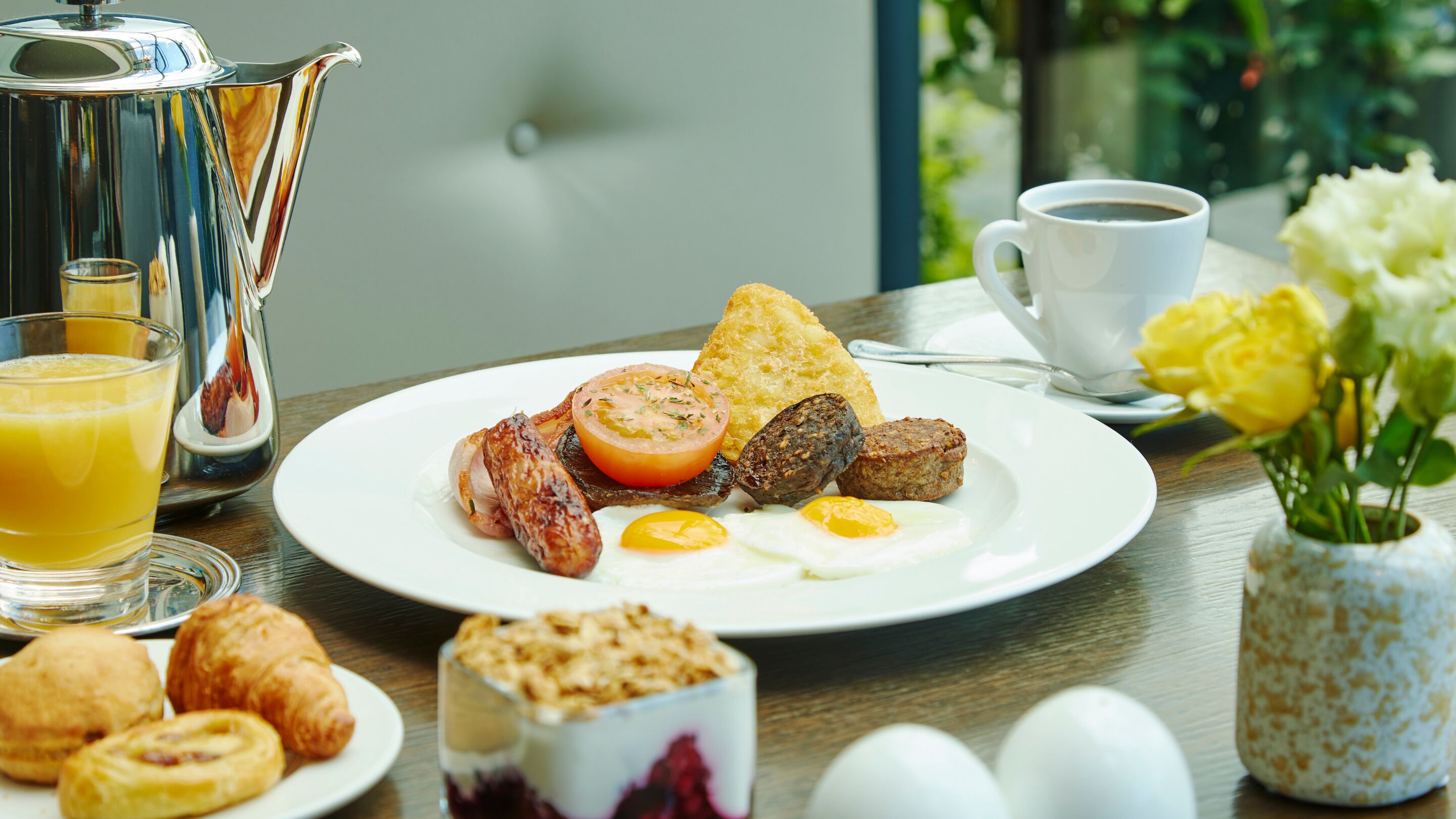 Ein Frühstückstisch mit Eiern, Würstchen und Toast, einem Glas Orangensaft, einer Kaffeetasse und einer Blumenvase.