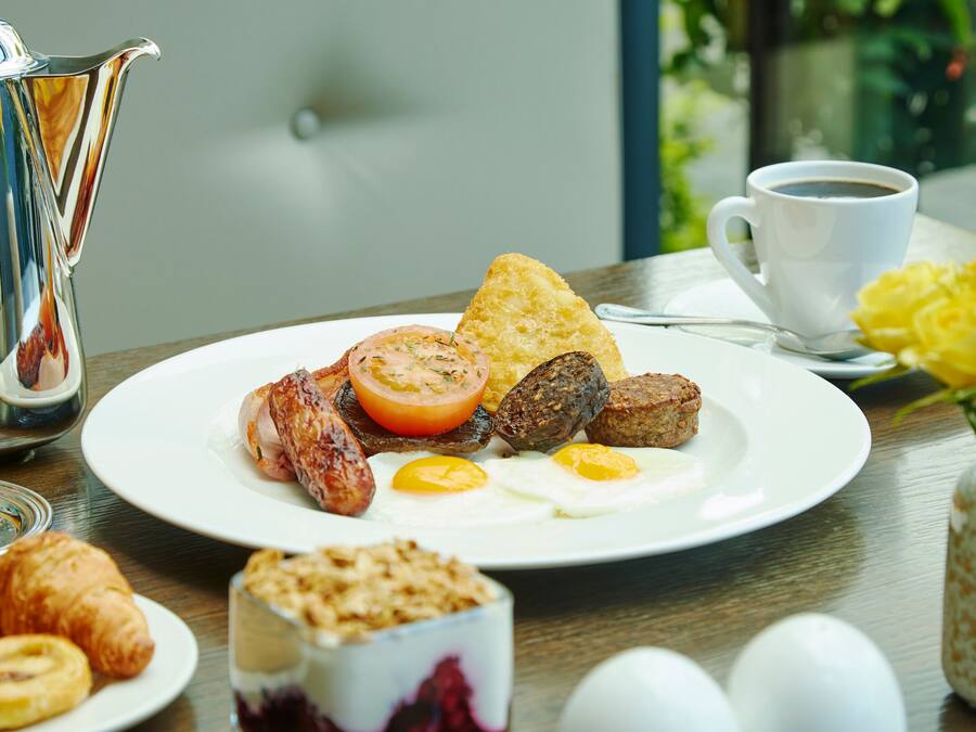Ein Frühstückstisch mit Eiern, Würstchen und Toast, einem Glas Orangensaft, einer Kaffeetasse und einer Blumenvase.