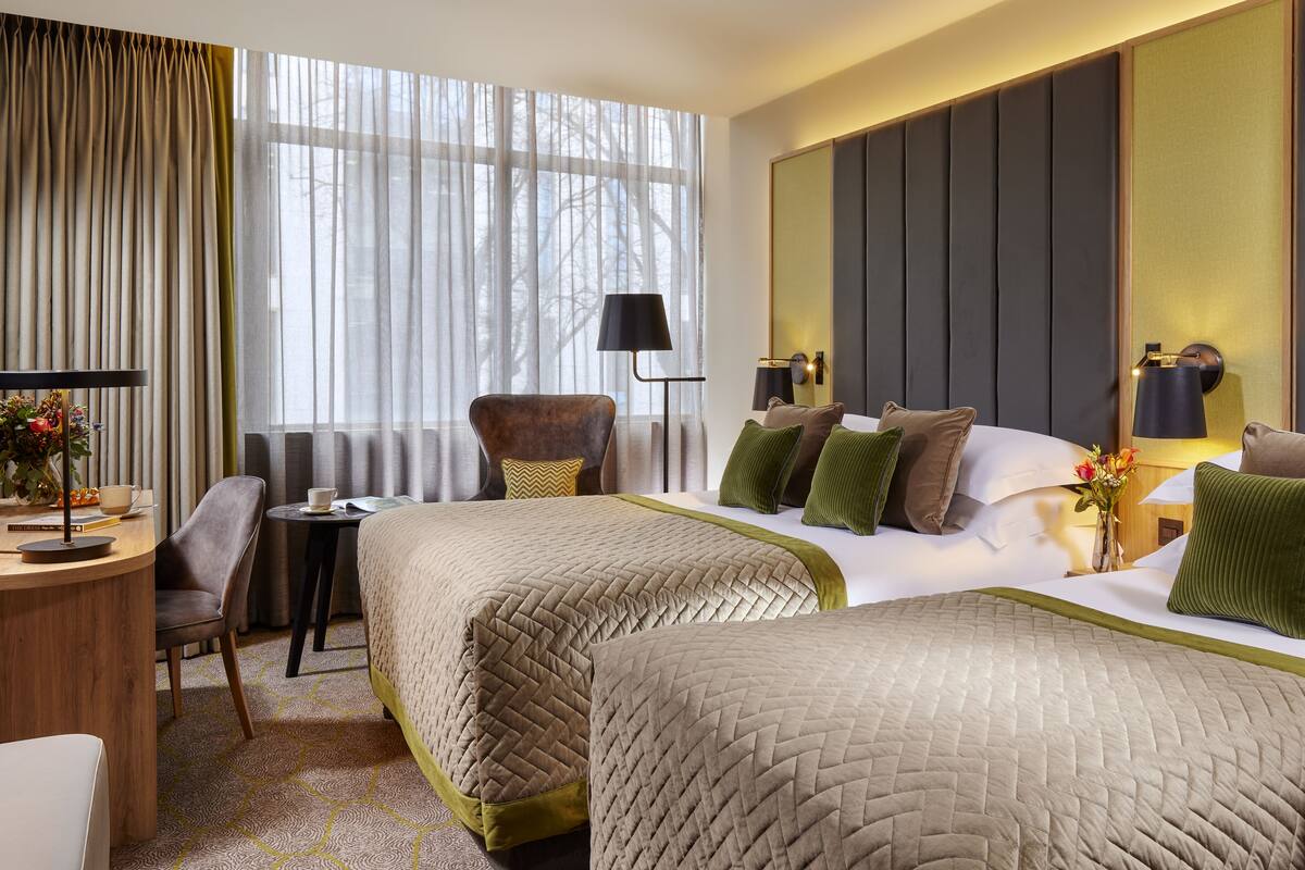 A hotel room with two beds, a desk, a chair, and a lamp.