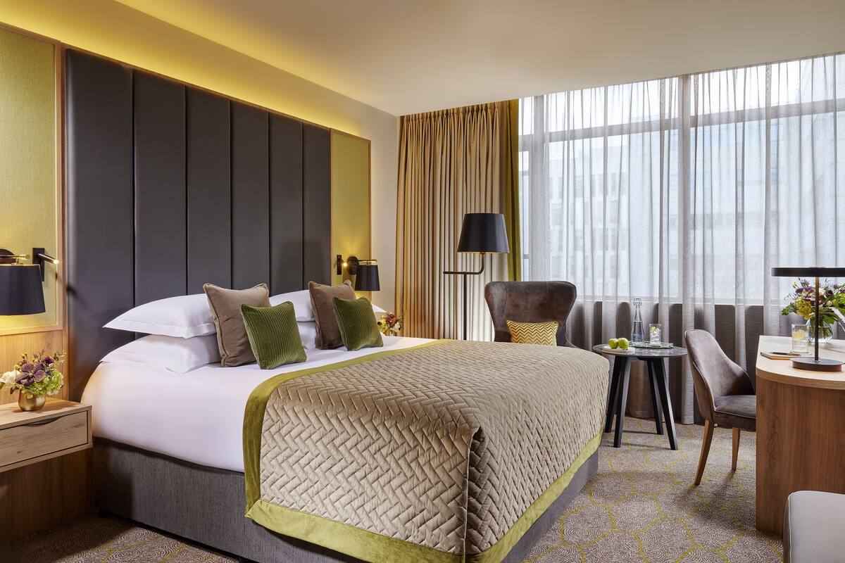 A hotel room with a large bed, a desk, a chair, and a view of the city through sheer curtains.