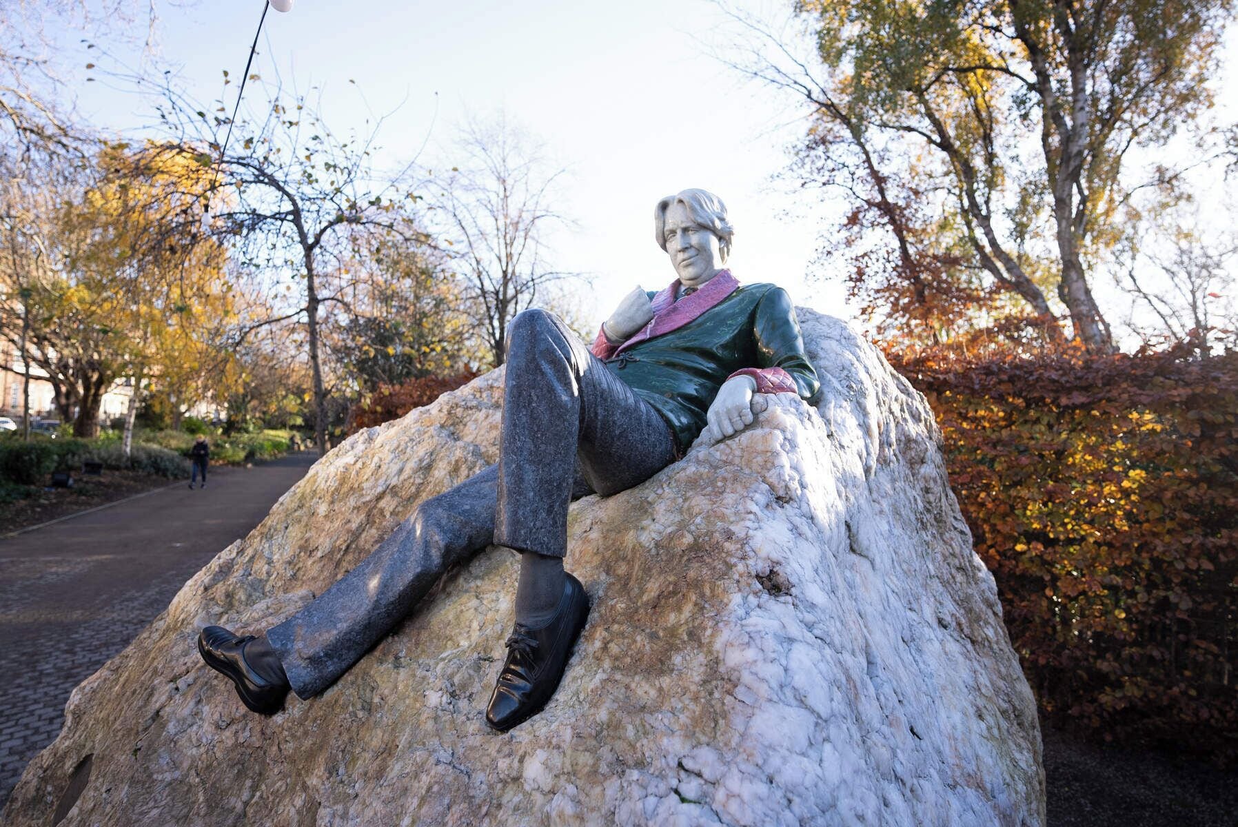 A statue of a person sitting on a large rock in an outdoor setting with trees and a pathway in the background.