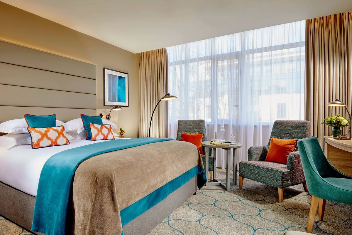 A hotel room with a large bed, two chairs, a small table, and a window with curtains.