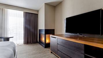Une chambre d’hôtel dotée d’une grande télévision à écran plat, d’une cheminée, d’un lit et offrant une vue sur la ville grâce à de grandes fenêtres.