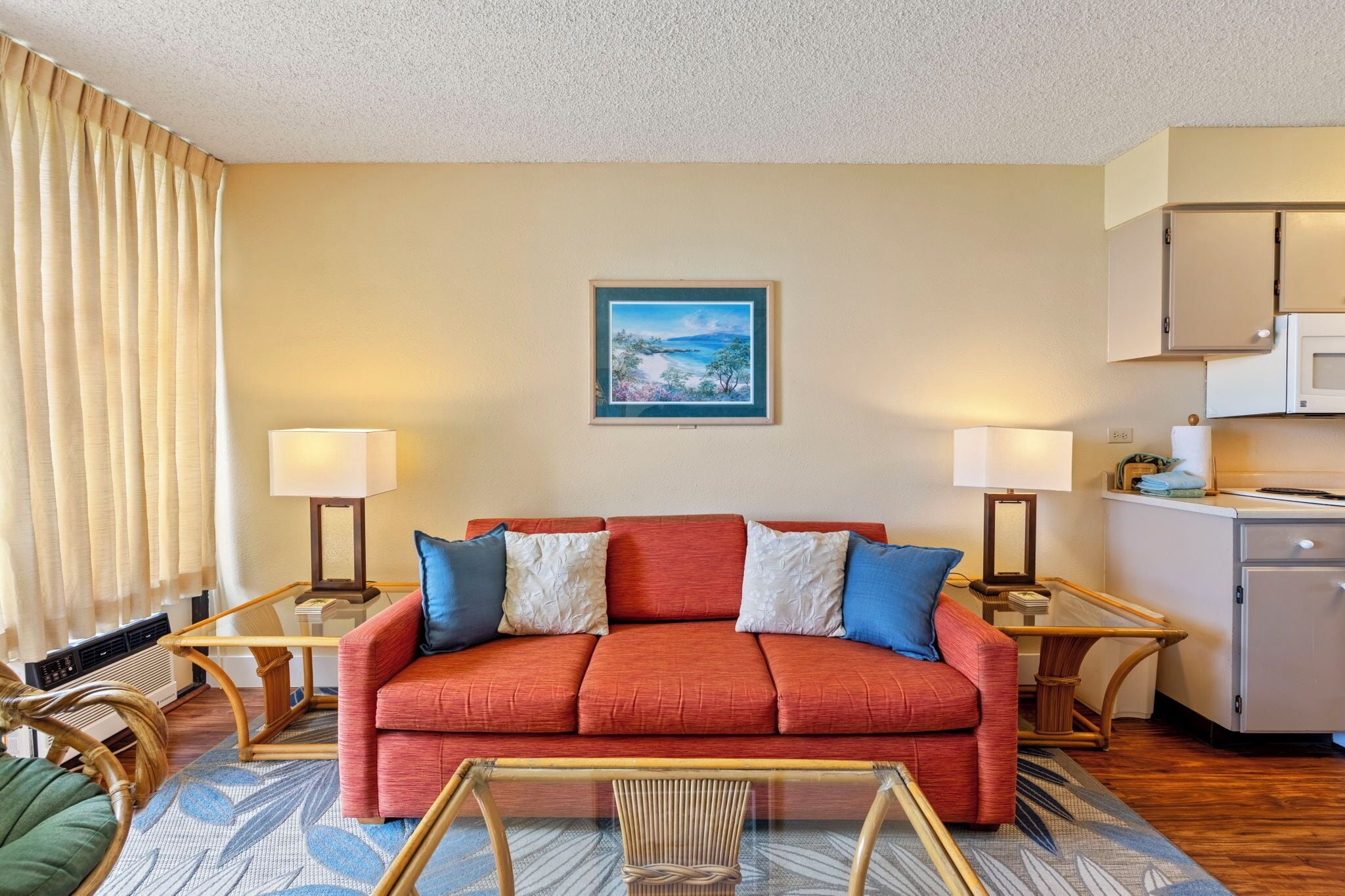A hotel room with a red sofa, two lamps, a framed picture on the wall, a kitchen area, and a window with curtains.