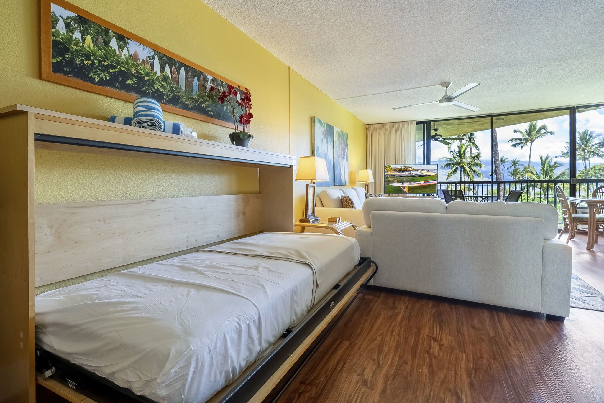Une chambre d’hôtel avec un lit, un canapé, un ventilateur de plafond et un balcon offrant une vue sur les palmiers.