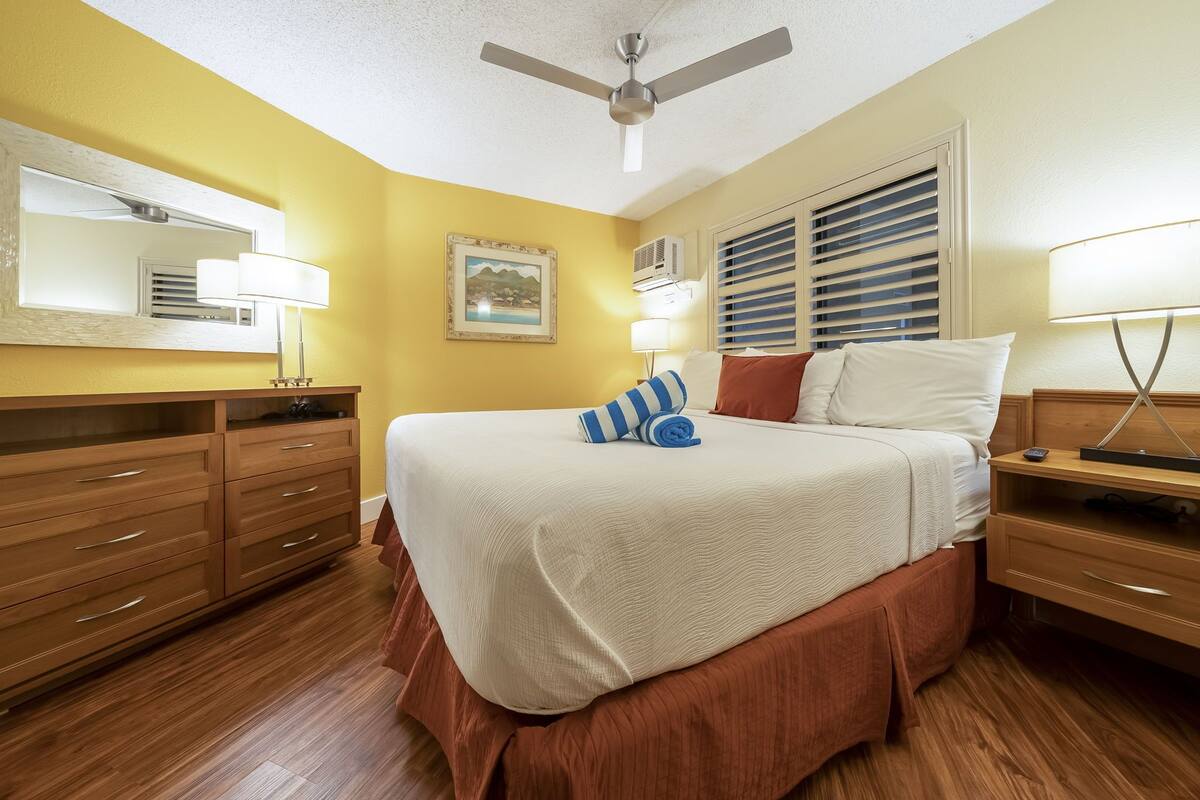 A hotel room with a bed, wooden dresser, ceiling fan, and a painting on the wall.