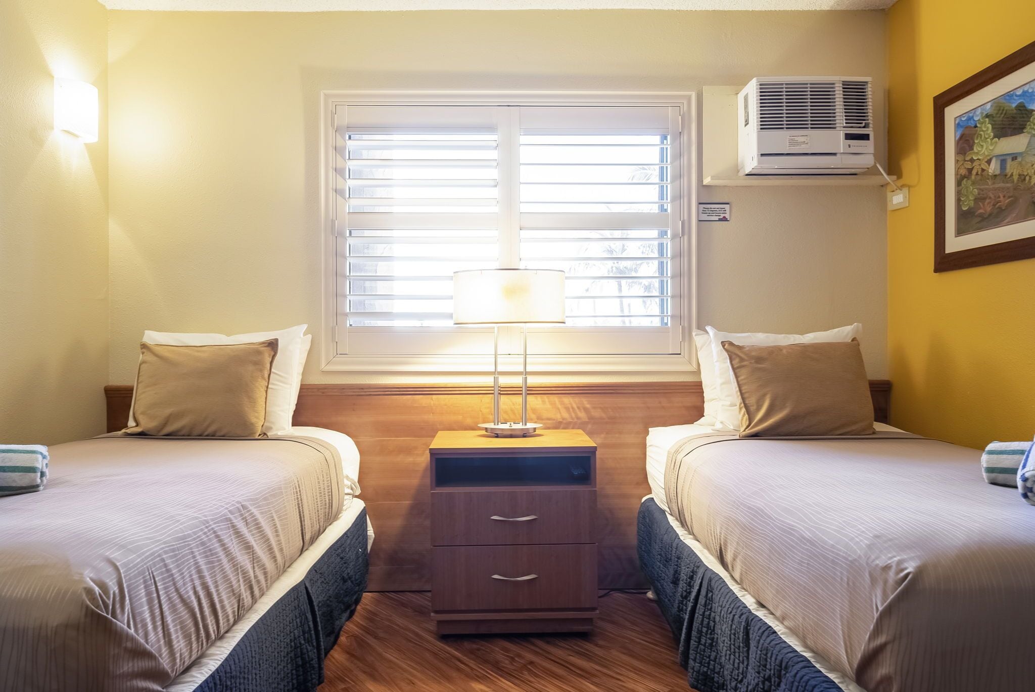A hotel room with two beds, a nightstand with drawers, a lamp, an air conditioning unit, and a window with blinds.