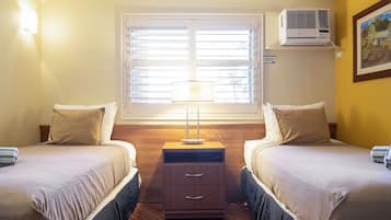 A hotel room with two beds, a nightstand with drawers, a lamp, an air conditioning unit, and a window with blinds.