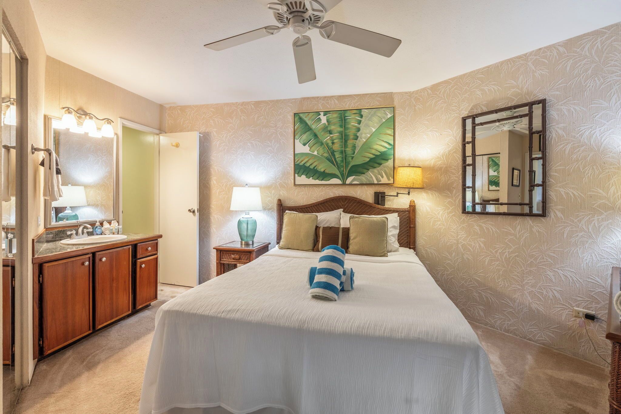 A hotel room with a bed, a ceiling fan, a large mirror, a wall-mounted lamp, and a bathroom with a sink and mirror.