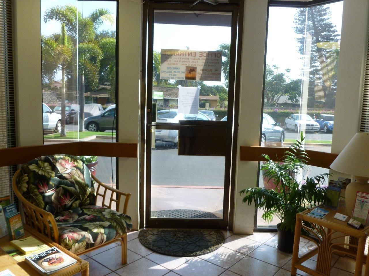 A hotel lobby with a tropical-themed chair, a wooden table, a lamp, and a large window offering a view of parked cars and palm trees outside.