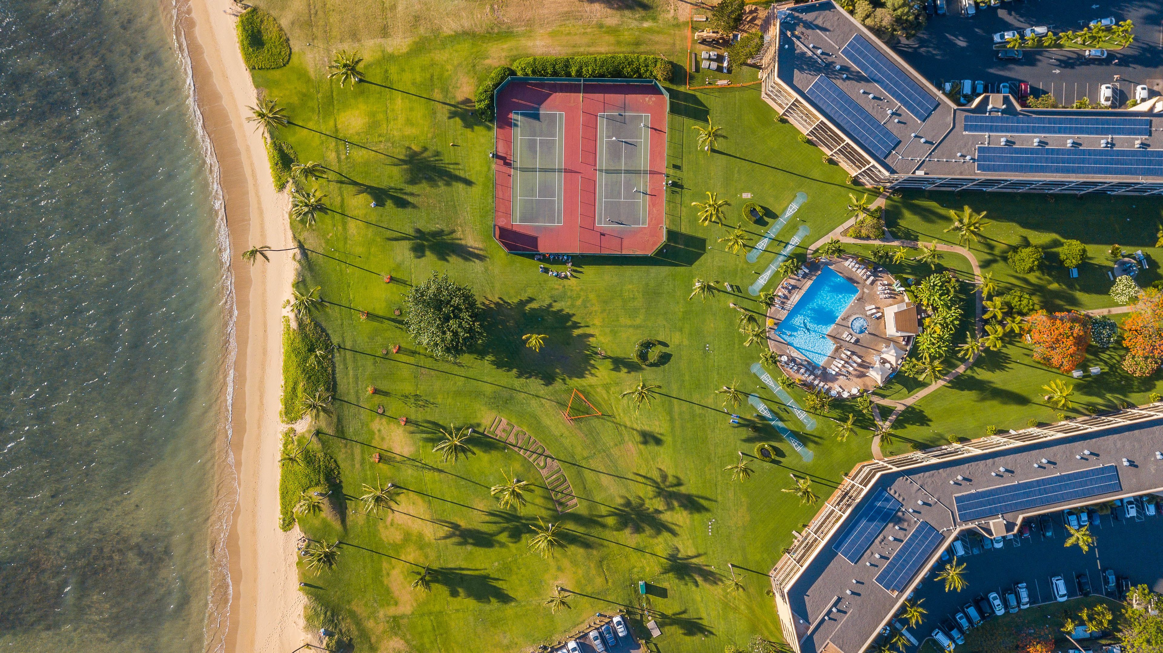 An aerial view of a resort with a tennis court, a swimming pool, and a beach.