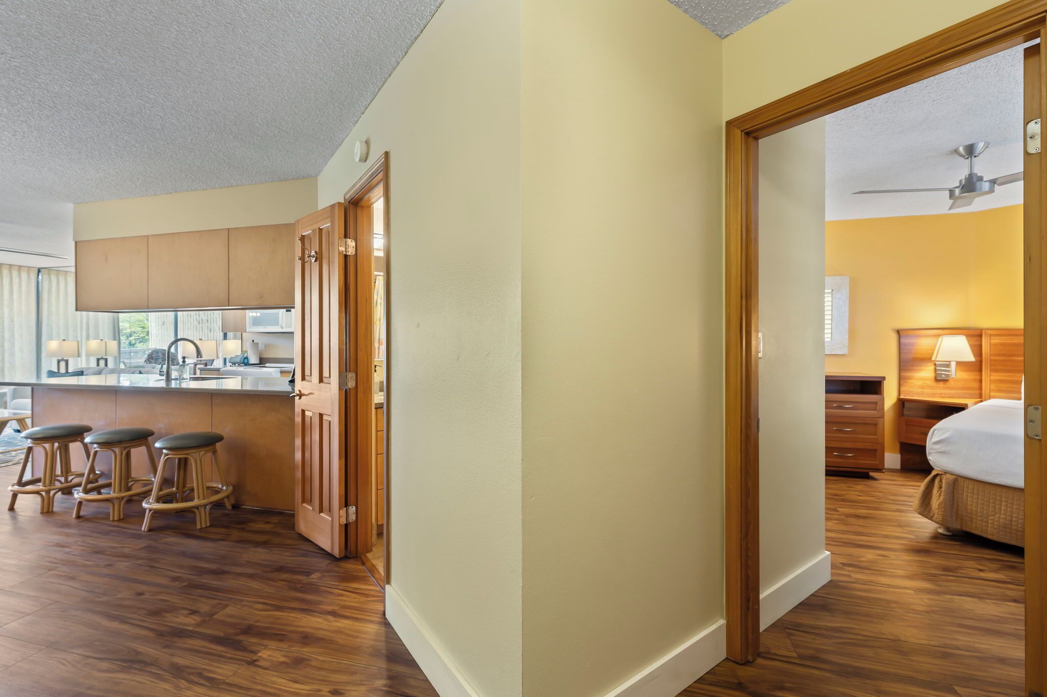 A modern kitchen with wooden cabinets, a bar area with stools, and a view of the living area with a bed and nightstand.