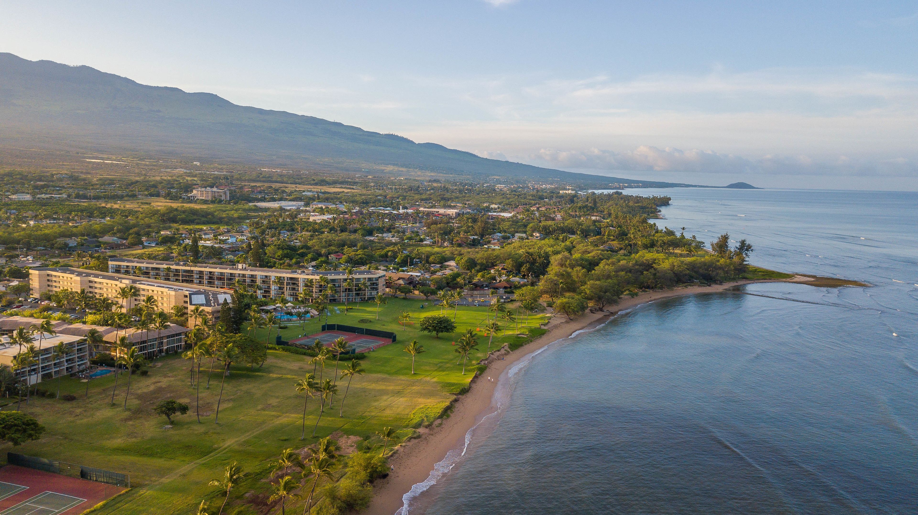 An aerial view of a coastal resort with multiple buildings, a beach, and a mountainous backdrop.