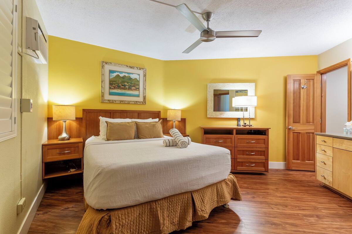 A hotel room with a bed, wooden nightstands, a ceiling fan, a painting on the wall, a mirror, and a wooden wardrobe.