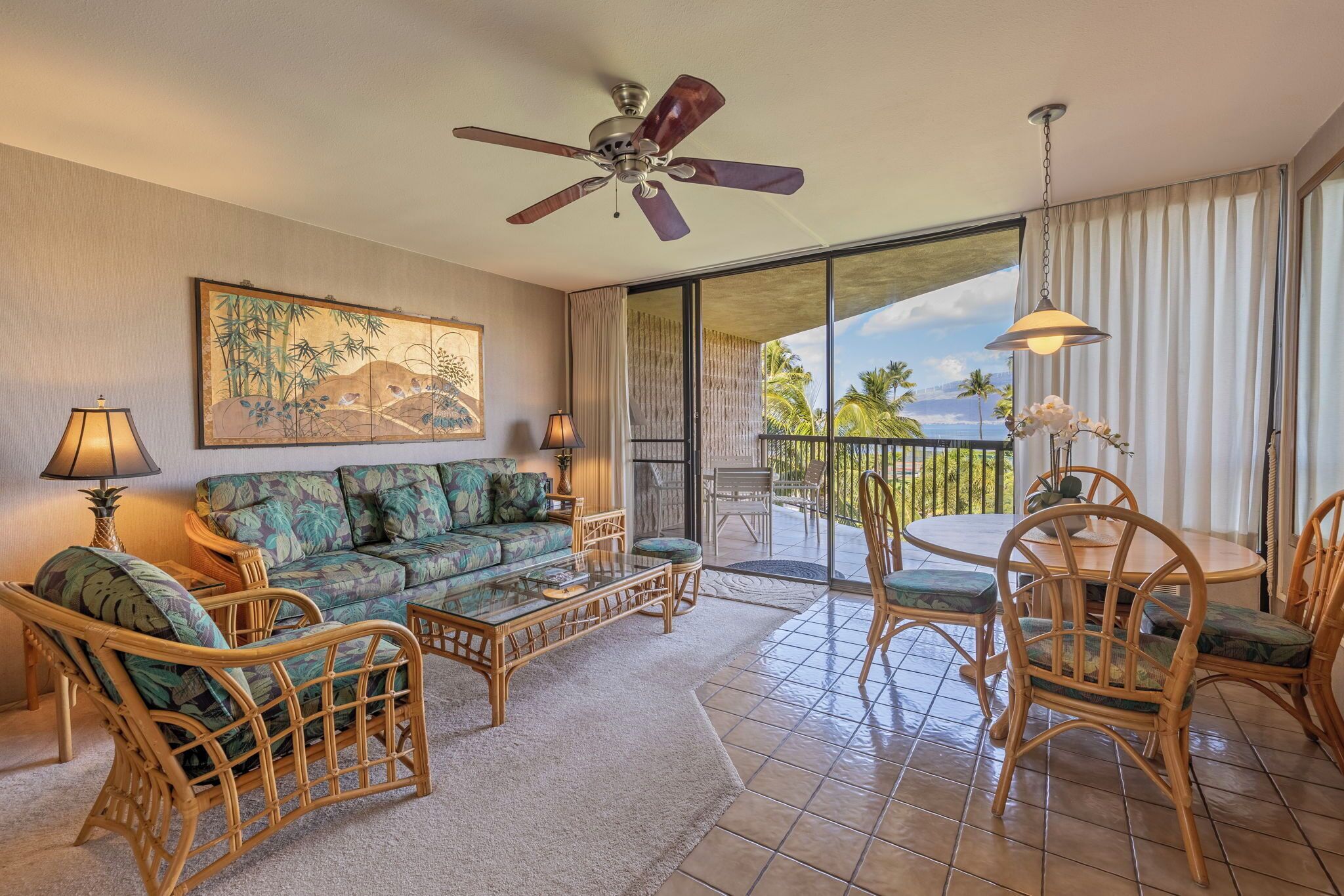A living room with a sofa, chairs, a coffee table, and a ceiling fan. There is a large window with a view of palm trees.