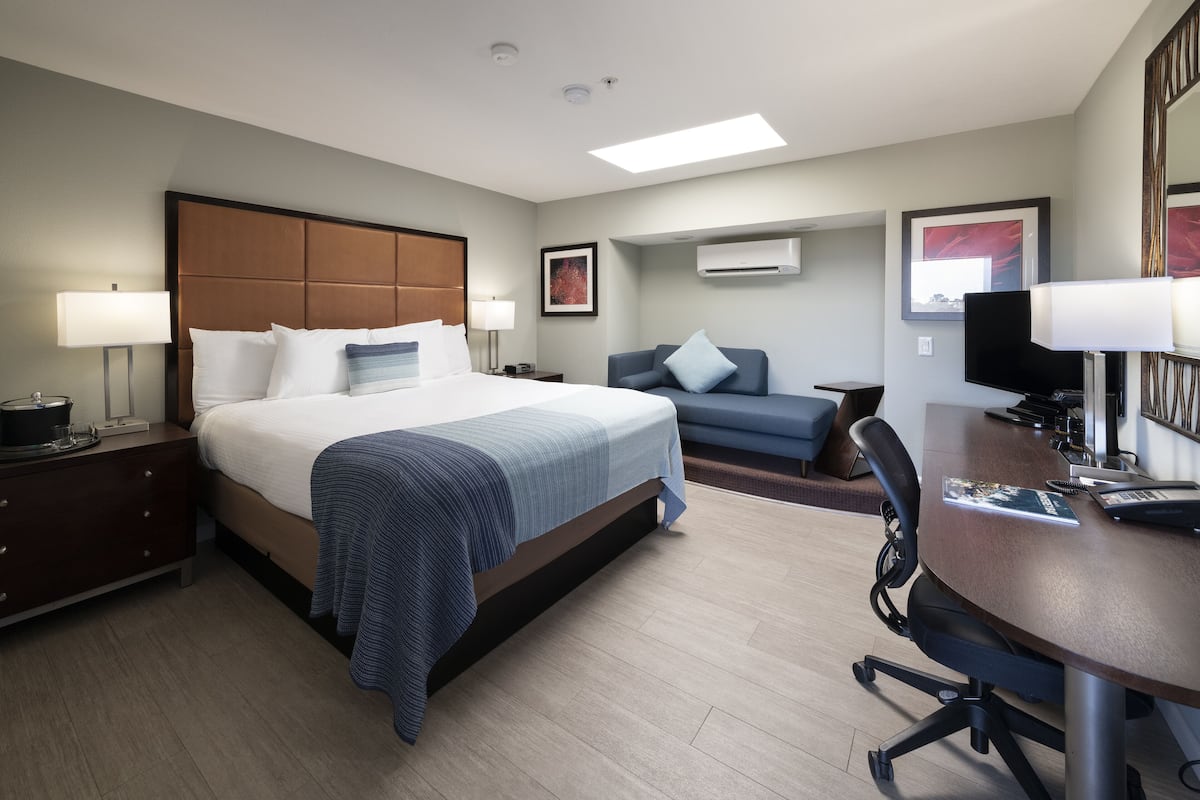 A hotel room with a large bed, a desk, a chair, a sofa, and an air conditioning unit.