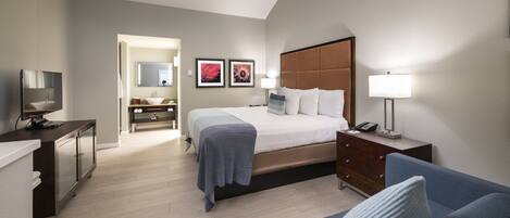 Une chambre d’hôtel moderne équipée d’un lit, d’un bureau, d’une télévision, d’un canapé et d’une salle de bain visible à travers une porte ouverte.