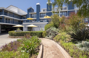 A hotel with a modern design, featuring balconies with yellow umbrellas, a landscaped garden with various plants, and a curved pathway leading to the entrance. - Mariposa Inn & Suites (Monterey)