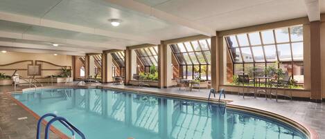 An indoor swimming pool with clear glass walls, a tiled floor, and a seating area with tables and chairs.