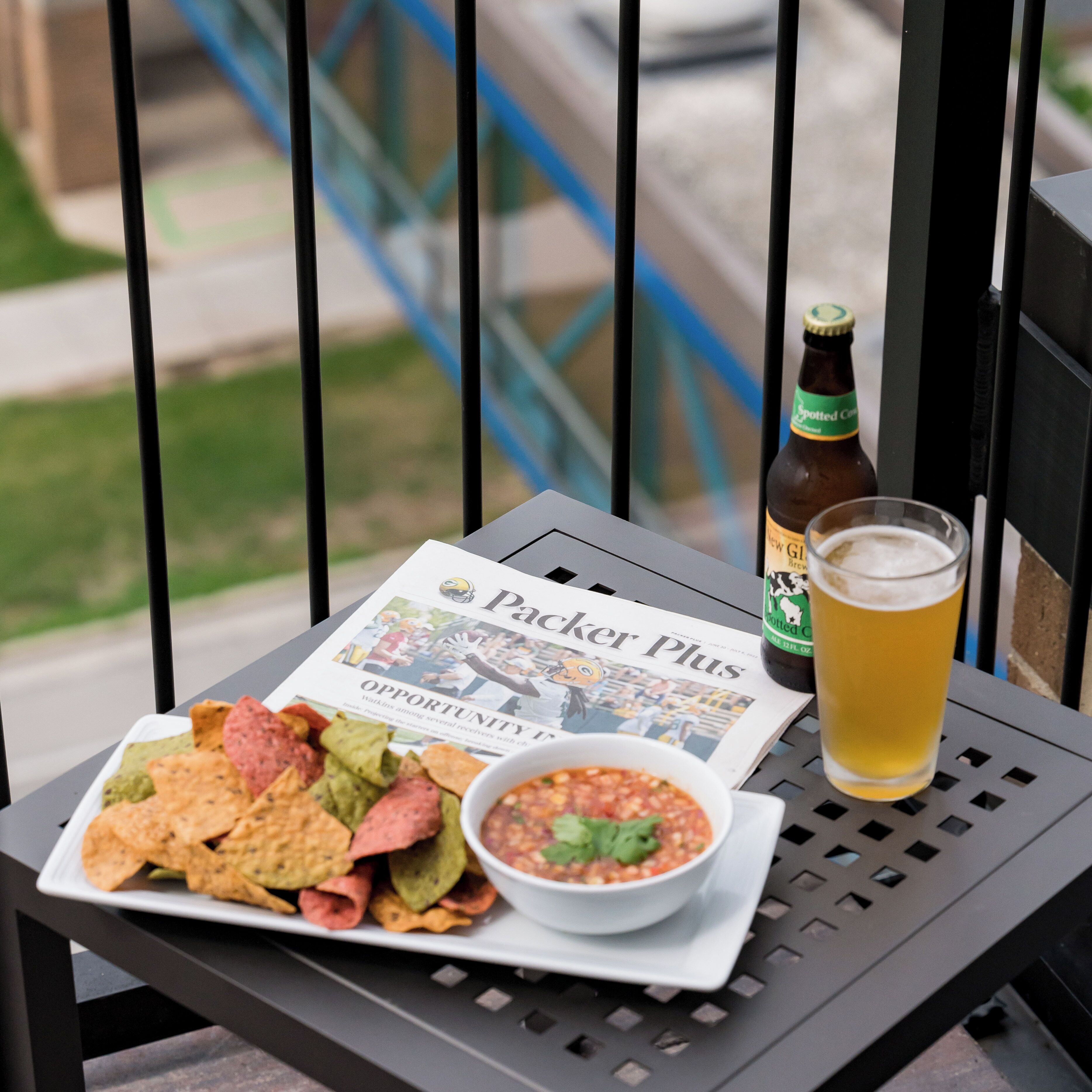 A table with a newspaper, a bottle of beer, a glass of beer, a bowl of salsa, and a plate of nachos.