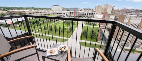 Un balcón con una mesa lista para el desayuno, una silla y vistas a un campus con edificios y zonas verdes.