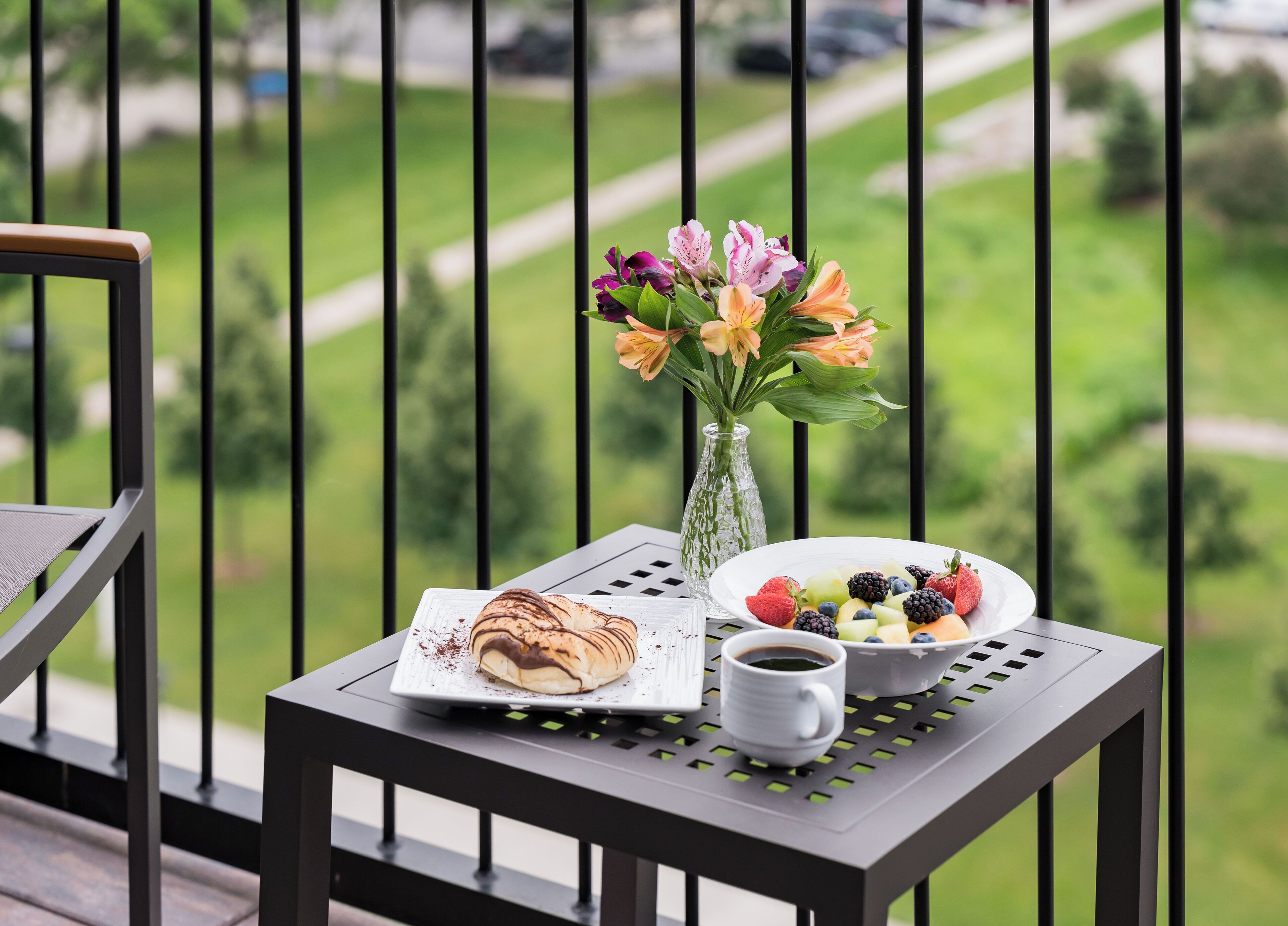 A balcony with a table set for breakfast, including a plate of fruit, a cup of coffee, and a pastry.