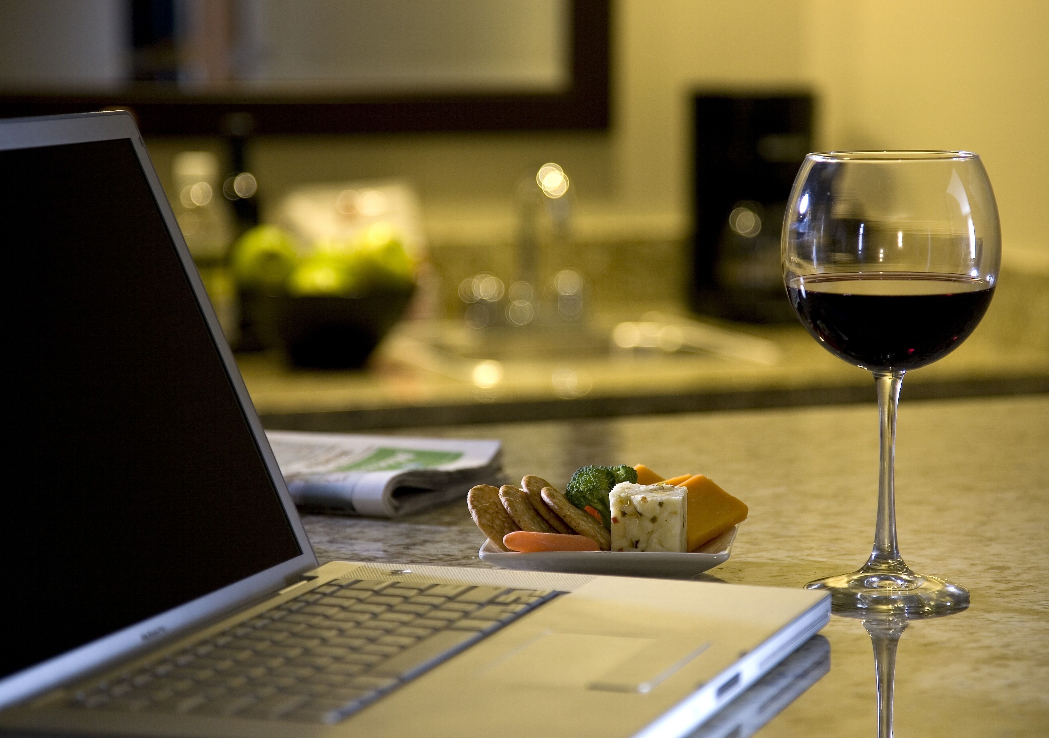 Una laptop, una copa de vino tinto y un plato con queso y galletas sobre una mesa.