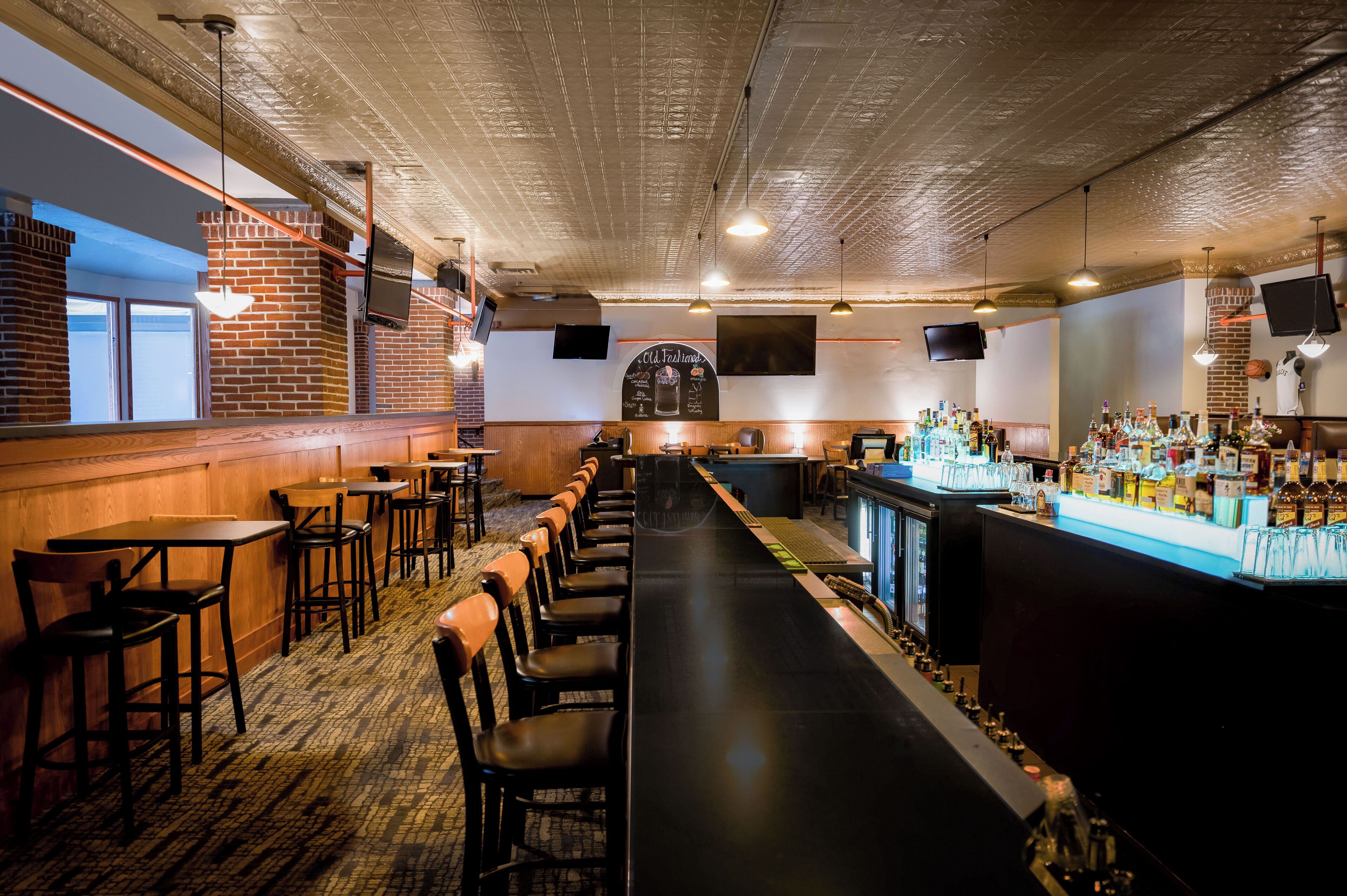 A bar with a long counter, wooden chairs, and a brick wall.