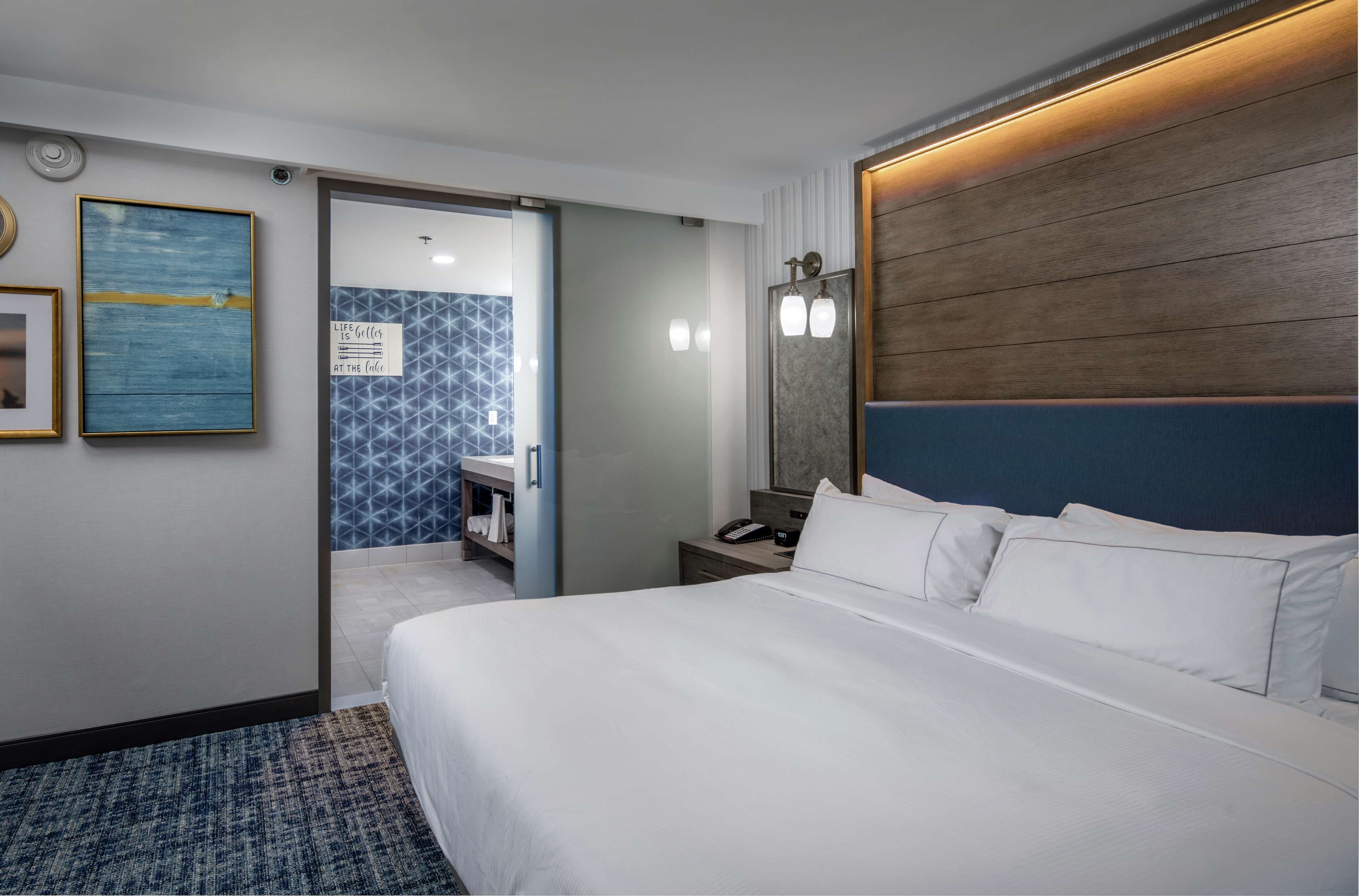 Une chambre d’hôtel avec un grand lit, une table de chevet avec une lampe, un miroir fixé au mur et une porte donnant sur une salle de bain équipée d’un porte-serviettes.