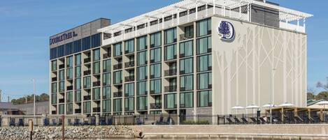 A modern hotel with a large glass facade, outdoor seating by the water, and a prominent logo on the side.