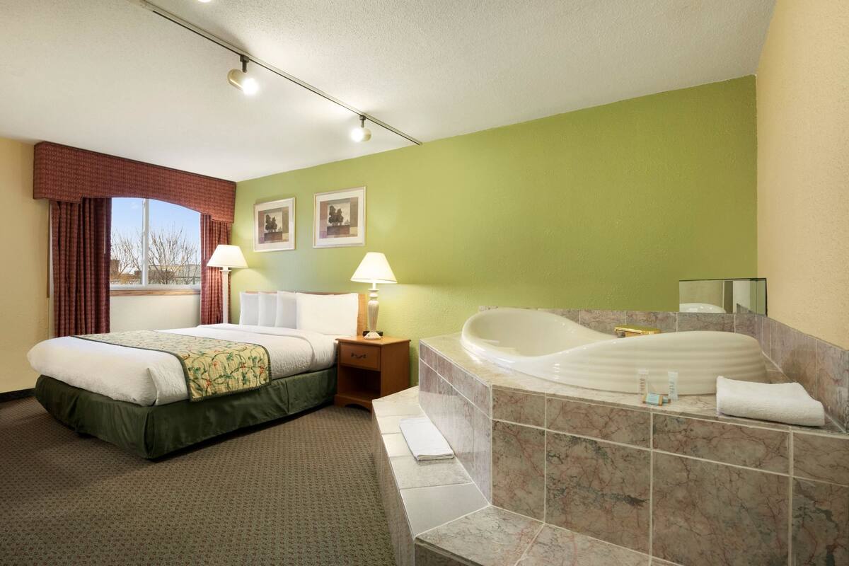 Habitación de hotel con cama, una bañera grande, una ventana con cortinas y dos lámparas fijadas a la pared.