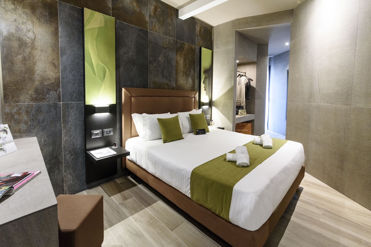 A modern hotel room with a large bed, a nightstand, a desk with a chair, and a mirror.
