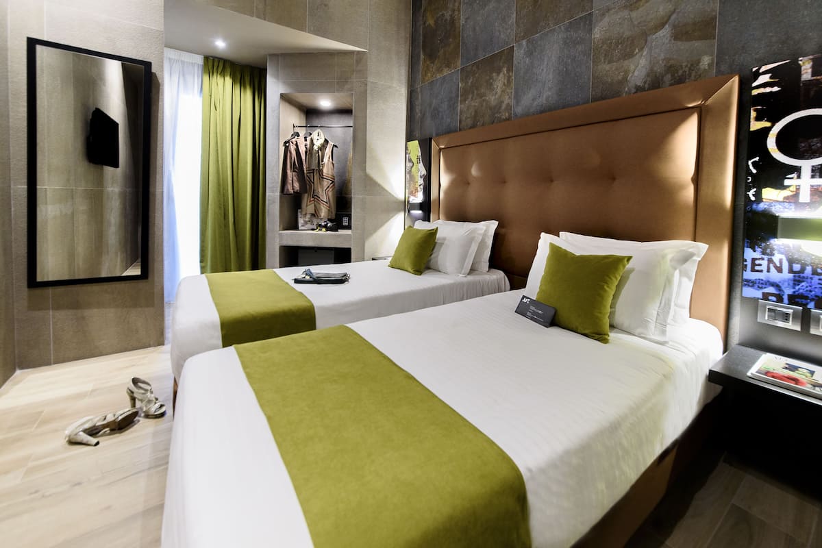 A hotel room with two beds, a mirror, a vanity with a mirror, and a coat rack with hanging clothes.