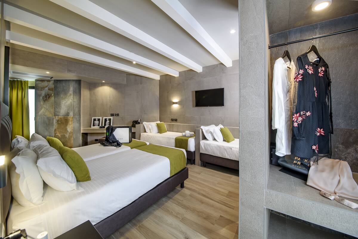 A hotel room with two beds, a flat-screen TV, a vanity with a mirror, and a clothing rack with dresses.