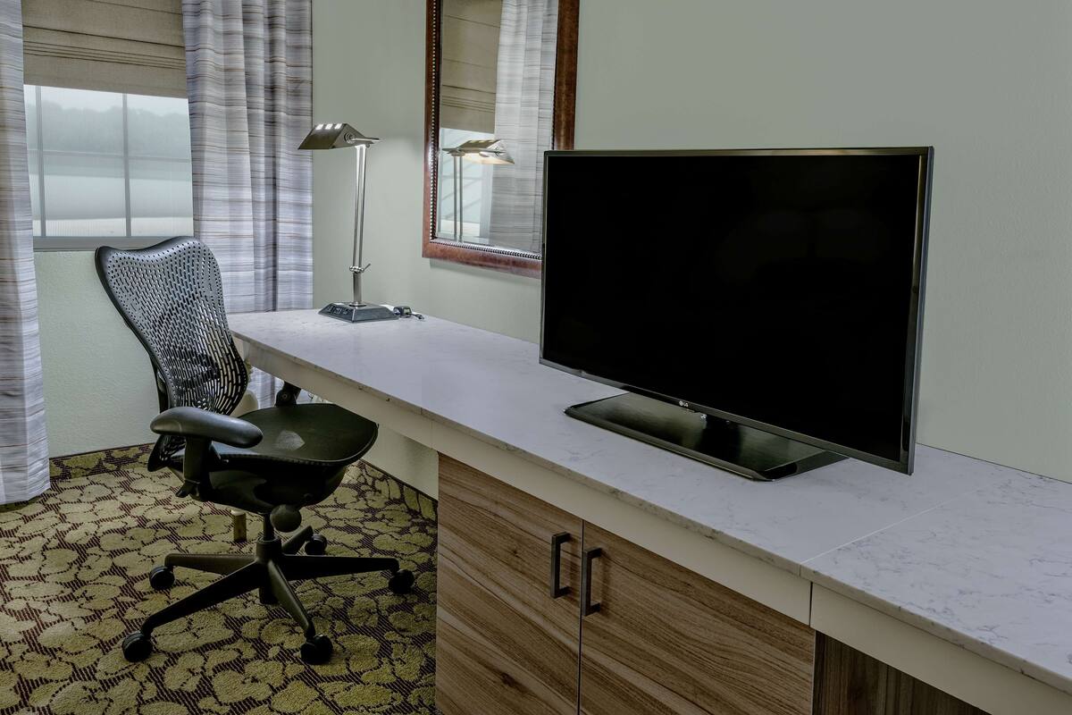 A hotel room with a large flat-screen TV, a desk with a lamp, a chair, and a patterned carpet.