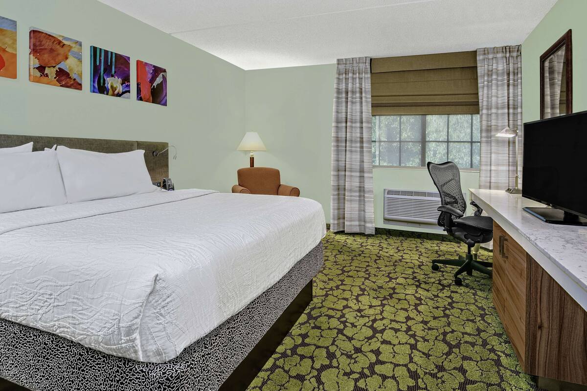 A hotel room with a large bed, a desk with a computer, a chair, a lamp, and a window with curtains.