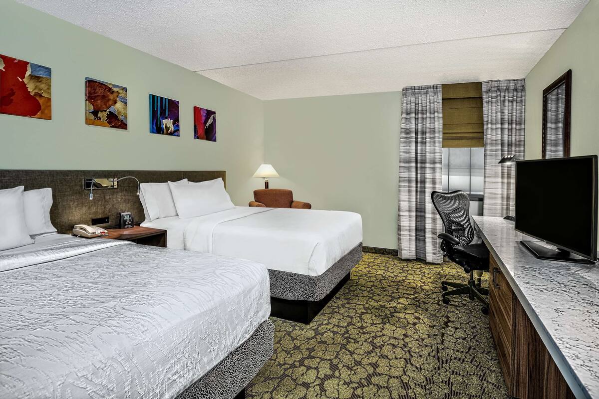 A hotel room with two beds, a desk with a computer, a lamp, and artwork on the wall.