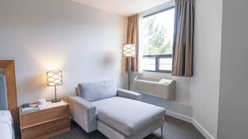 Una habitación de hotel moderna con un sofá gris, una mesita de noche de madera con lámpara y una ventana con cortinas.