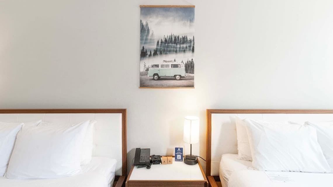 Habitación de hotel con dos camas, una mesita de noche con teléfono, una lámpara y un cuadro de una furgoneta en un bosque.