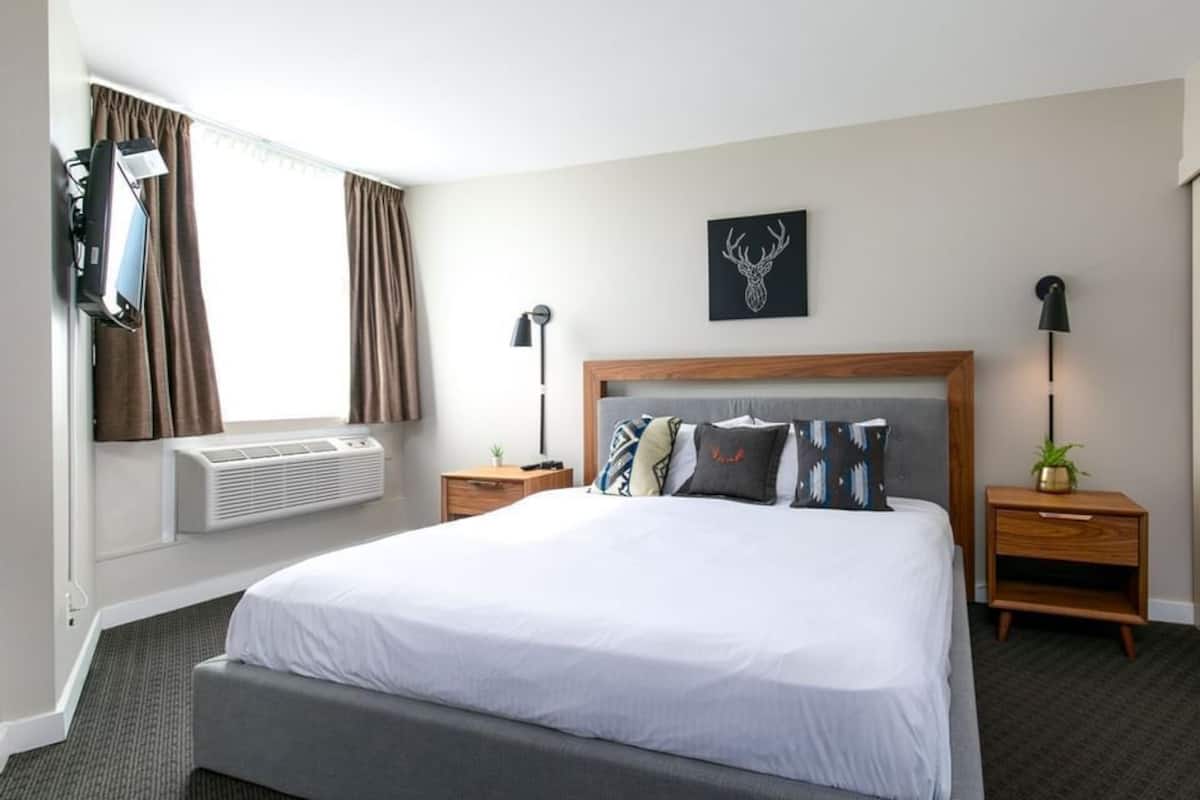 A hotel room with a large bed, two bedside tables, a wall-mounted TV, a ceiling fan, and a window with curtains.