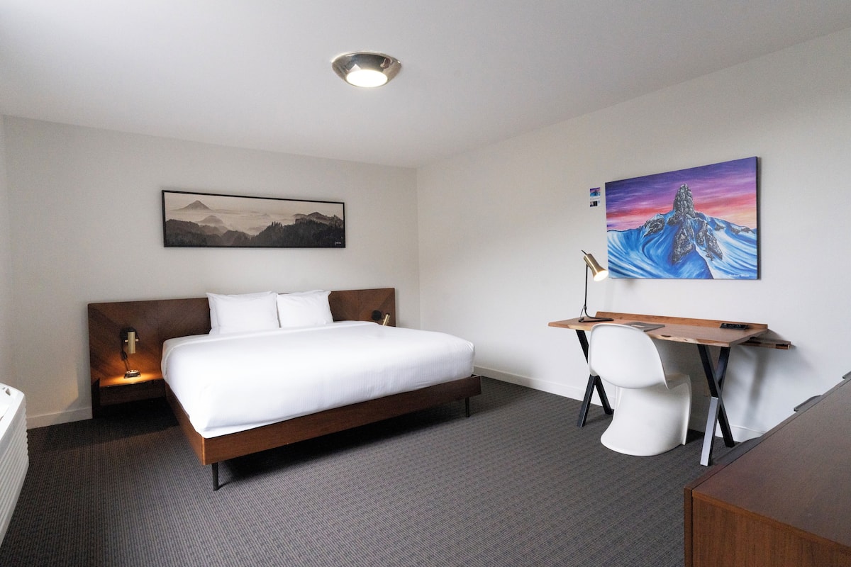 A hotel room with a bed, desk, and chair, a painting on the wall, and a lamp on the nightstand.