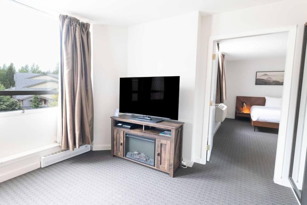 A hotel room with a large flat-screen TV on a wooden stand, a bed with a headboard, and a window with curtains.