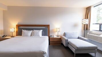 A hotel room with a large bed, bedside tables with lamps, a grey armchair, and a window with curtains.