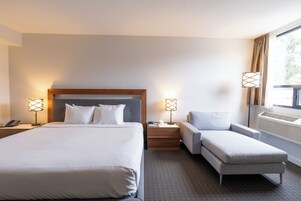 A hotel room with a large bed, bedside tables with lamps, a grey armchair, and a window with curtains.