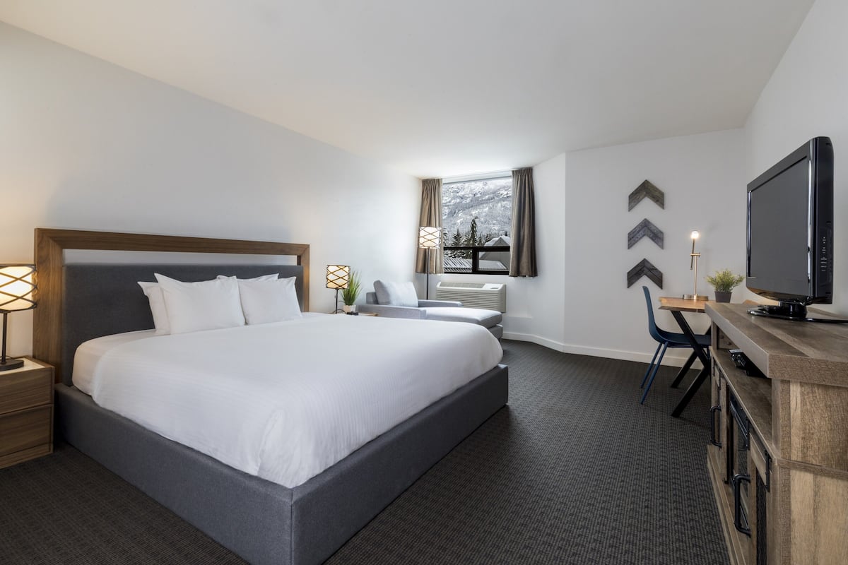 A modern hotel room with a large bed, bedside tables with lamps, a desk with a chair, a television, and a window with a view of a snowy landscape.