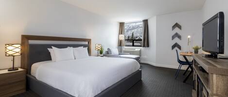 Une chambre d’hôtel moderne avec un grand lit, des tables de chevet avec des lampes, un bureau avec une chaise, une télévision et une fenêtre donnant sur un paysage enneigé.