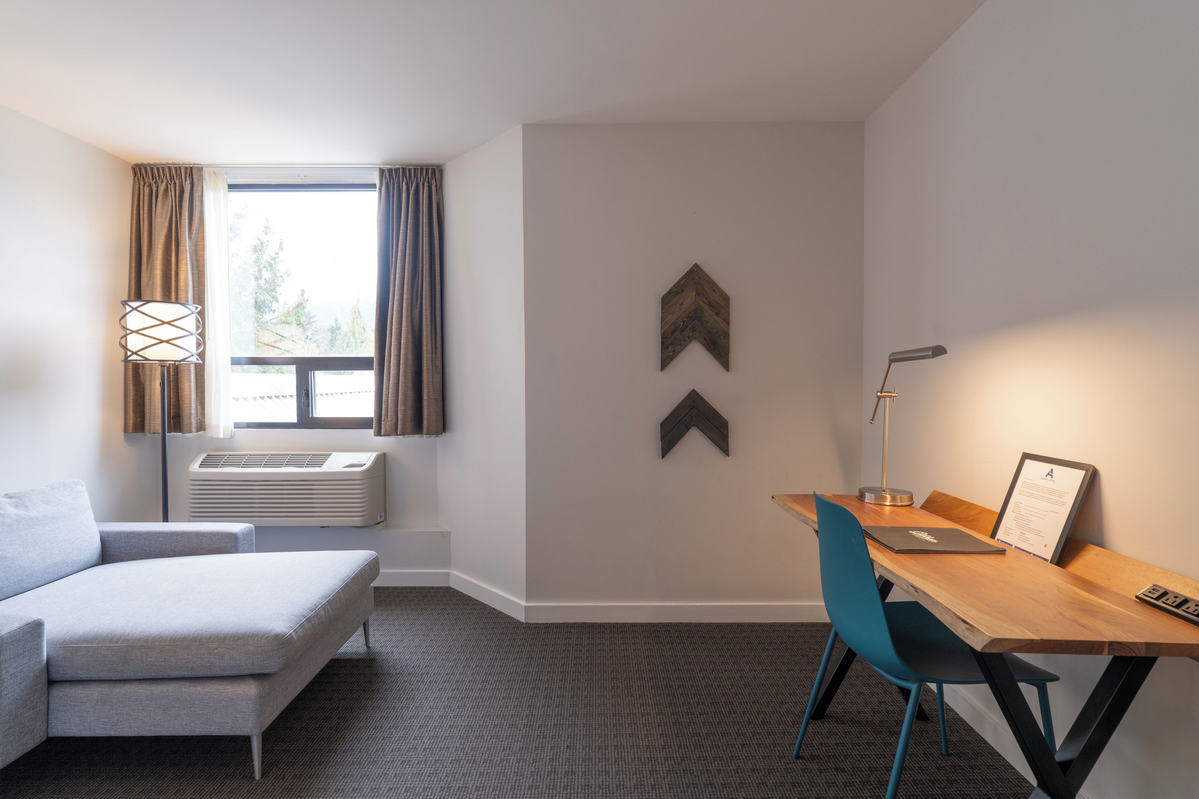 A modern hotel room with a grey sofa, a wooden desk with a laptop, a blue chair, and a large window with curtains.